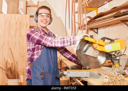 Giovane donna come un falegname apprendista con protezione auricolare funziona con la troncatrice Foto Stock