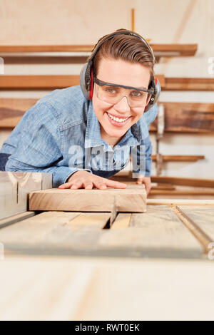 La donna come un falegname apprendista con occhiali protettivi e una protezione per le orecchie seghe sul legno della sega circolare Foto Stock