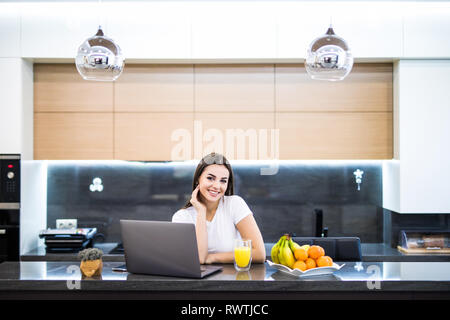 L'immagine verticale di una giovane donna sorridente seduto davanti al suo computer in cucina Foto Stock