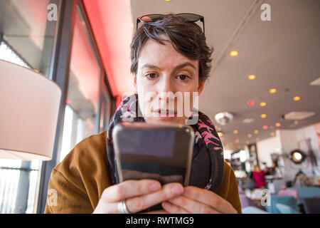 Bella giovane donna in abiti caldi in seduta cafe e controllando il suo smartphone, sorpreso e confuso, sfondo sfocato, testa e spalle
