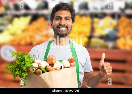 Amichevole maschio indiano il supermercato o ipermercato lavoratore titolare carta marrone sacchetto shopper con il pollice in alto come gesto Foto Stock