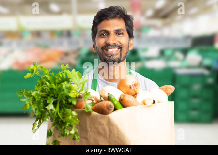 Maschio indiano drogheria o ipermercato lavoratore titolare carta marrone borsa di verdura con un sorriso amichevole espressione Foto Stock