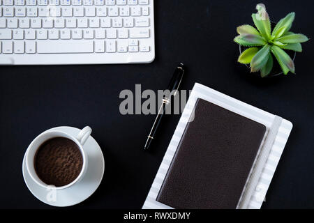 Office desk top view o laici piatta con sfondo nero, bianco tastiera, tazza di caffè, notebook con la penna nera e una piccola succulenta. Foto Stock