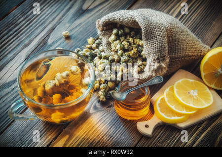 Una sana camomilla tazza da tè, hessian sacchetto di margherite secchi fiori, miele il vaso e le fette di limone sul bordo di taglio. Foto Stock