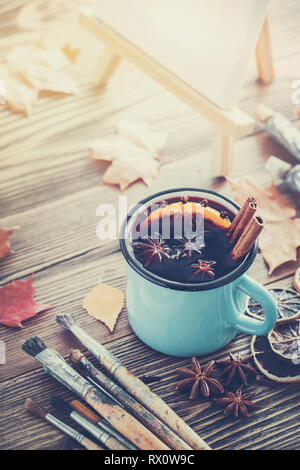 Tazza di vin brulé, artista pennelli, vernici, tela artistica sul cavalletto e foglie di autunno sulla scrivania. Foto Stock