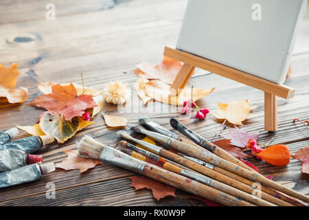 Tela su cavalletto, tubi di vernice, pennelli per dipingere e foglie di autunno sul vecchio sfondo di legno. Retrò tonica. Foto Stock