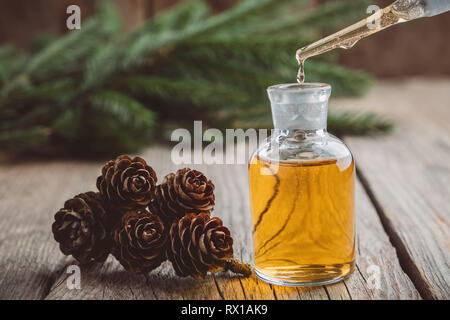 Fir olio aromatico in un vetro flacone contagocce, una goccia di essenziali di pino gocciolamento di olio dal vetro contagocce. Coni di abete, di conifere, rami di alberi in backgr Foto Stock