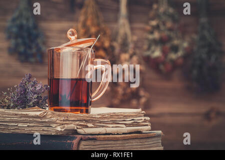 Tazza di tè sano o tintura a base di erbe sulla pila di libri antichi. Appendere i grappoli di erbe medicinali su sfondo. La medicina di erbe. Foto Stock