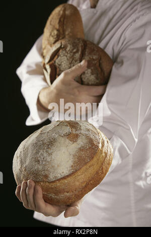 Baker in bianco uniforme che porta fresca Pane di segale pani nelle sue mani, offrendo uno verso la telecamera in close-up, in piedi su sfondo nero Foto Stock