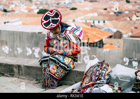 Una Donna vestita di tradizionale Peruviano abbigliamento siede su un banco di pietra mentre la realizzazione e la vendita di regali fatti a mano (Cusco, Perù) Foto Stock