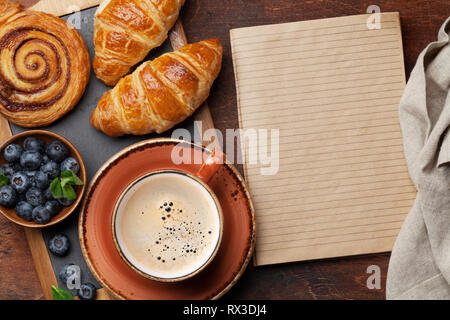 Caffè, cornetti, panini alla cannella e le bacche di prima colazione. Su un tavolo di legno. Con il blocco note per il testo Foto Stock