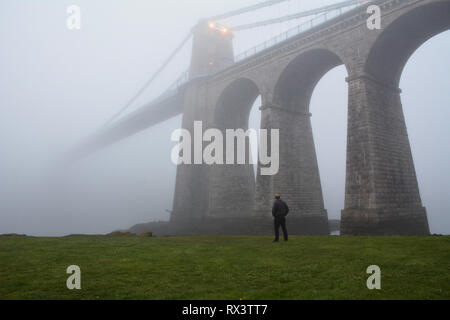 Osservando il Menai Suspension Bridge nella nebbia e la nebbia che scompare oltre verso Anglesey. Unire la terraferma gallese di Anglesey. Foto Stock