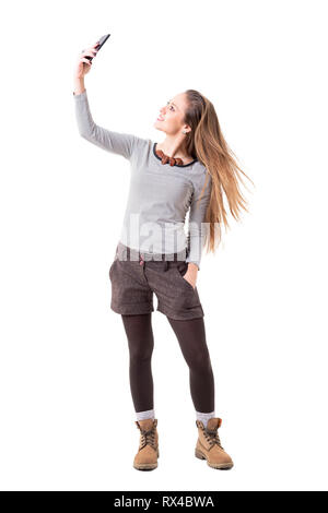 Felice eccitato giovane ragazza hipster tenendo selfie e lanciando che scorre lungo i capelli biondi. Corpo pieno isolati su sfondo bianco. Foto Stock