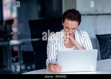 Un giovane imprenditrice siede dietro il suo computer portatile accigliata con la sua testa con le mani a causa di un problema al lavoro Foto Stock