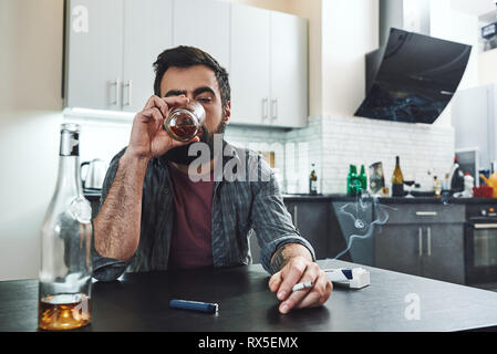 Dai capelli scuri, triste e sprechi di gradazione uomo seduto al tavolo, in cucina, fumare e bere whisky, tenendo il vetro, completamente ubriaco, cercando Foto Stock