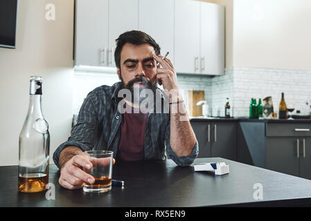 Dai capelli scuri, triste e sprechi di gradazione uomo seduto al tavolo, in cucina, fumare e bere whisky, tenendo il vetro, completamente ubriaco, cercando Foto Stock