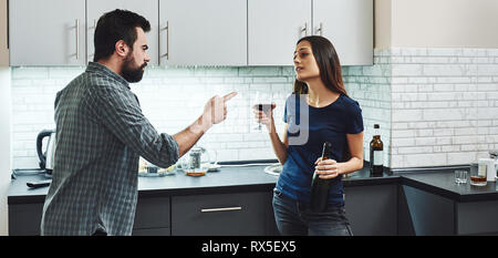 Ubriaco. Dai capelli scuri addicted donna litigare con suo marito. Ella è in piedi in cucina con un bicchiere e una bottiglia di vino rosso e ascolto di h Foto Stock