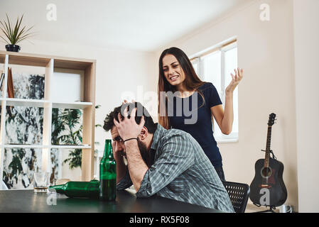 Bevuto il marito. Dai capelli scuri addicted sentimento donna disperazione mentre gridava a suo marito ubriaco seduta in cucina Foto Stock