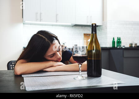 Dopo aver bevuto. Assonnato bevuto giovane donna giaceva sul tavolo in cucina e di dormire con la bottiglia e bicchiere di vino rosso in piedi sulla tavola in fro Foto Stock