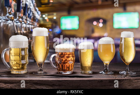 Refrigerate tazze e bicchieri di birra sul vecchio tavolo in legno. Pub interno e banco bar con birra rubinetti a sfondo. Assortimento di birra. Foto Stock