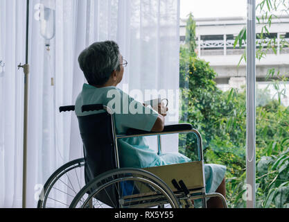 Senior uomo in una sedia a rotelle da solo in una camera guardando attraverso la finestra dell'ospedale. Paziente anziano Foto Stock
