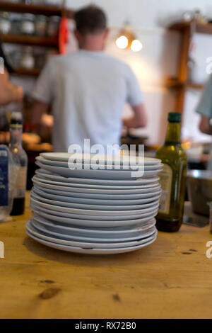 Una pila di piatti bianchi scheggiati si siede su un tavolo di legno di pino in primo piano in cucina con tre uomini che cucinano in background Foto Stock