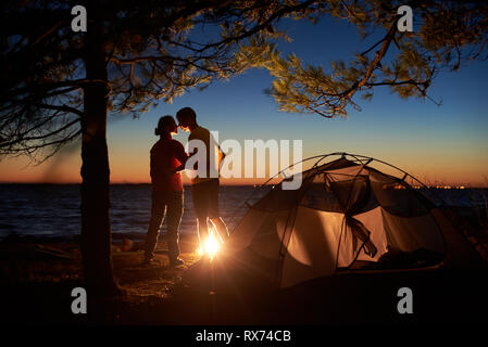 Notte Campeggio sotto agli alberi in mare. Sagome di escursionista giovane, vista posteriore di un uomo e di una donna stand al falò, abbracciando vicino a tourist tenda in serata. A Foto Stock
