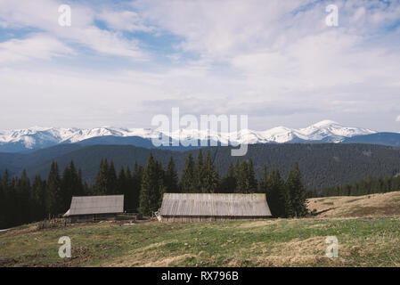 La molla paesaggio con montagne di neve. Case di legno di pastori sul pendio di una collina. Giornata di sole con nuvole. Bosco di abeti sulle colline. Karpaty, Ukrain Foto Stock