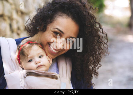 Close-up ritratto di una giovane madre attraente che porta un bambino in un vettore imbracatura mentre passeggiate all'aperto Foto Stock