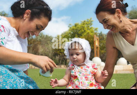 Una famiglia felice di 3 generazioni, baby, madre e nonna seduta sul prato e giocare all'aperto in un parco pubblico Foto Stock