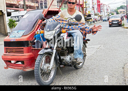 Roxas City, Capiz Provincia, Filippine: a basso angolo di visione del passaggio di un triciclo con un povero cercando metà età saluto del driver Foto Stock