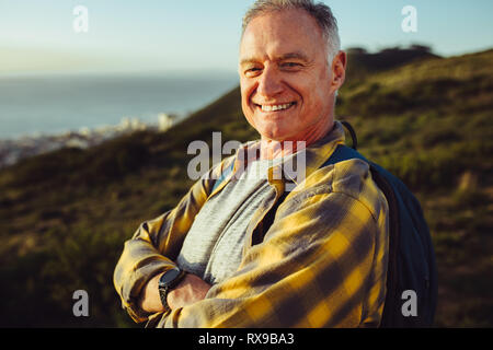 Close up ritratto di un anziano uomo in piedi all'aperto indossando uno zaino. Vista laterale di un allegro uomo in piedi su una collina con le braccia incrociate. Foto Stock