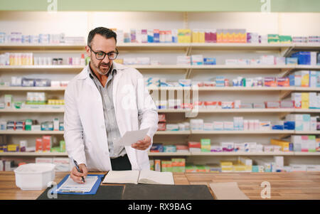 Il farmacista maschio prescrizione di scrittura al posto di lavoro. Maschio maturo farmacista la lettura di una carta di prescrizione e la scrittura su appunti al contatore. Foto Stock