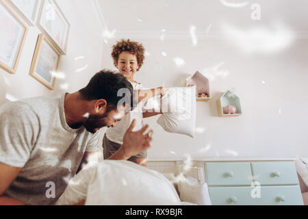 Happy boy cuscino in lotta con suo padre a casa. Padre e figlio avente una lotta di cuscini sul letto di piume battenti intorno a. Foto Stock