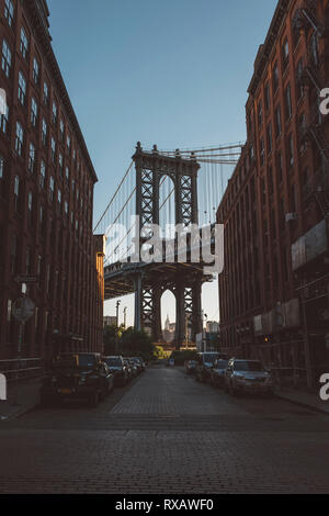 Auto parcheggiata su strada in mezzo edifici contro Manhattan Bridge durante il tramonto Foto Stock