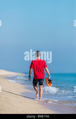 Vista posteriore dell'uomo azienda sport scarpe camminando sulla spiaggia a spiaggia contro sky Foto Stock