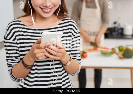 Ritratto di un simpatico giovane donna nelle cuffie che indossa il grembiule tramite cellulare per la cucina Foto Stock