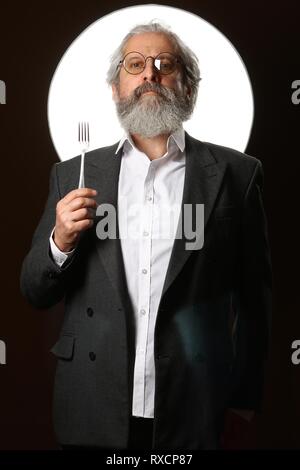 Ritratto in studio di un colore grigio-dai capelli uomo anziano con un shaggy i baffi e barba, in una camicia bianca e giacca grigia, indossare occhiali e una forcella su blac Foto Stock