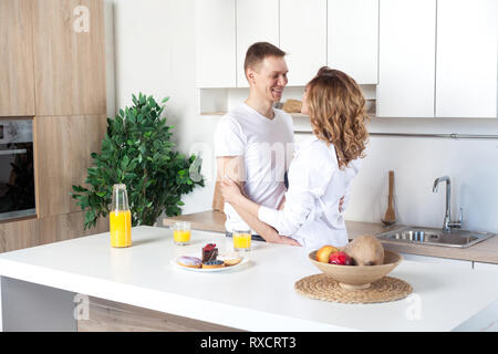 Giovani felice coppia sposata abbraccia in piedi vicino a tavola in cucina. Il marito abbraccia la sua moglie incinta, ai genitori di godere di ogni altro guardando agli occhi. Amore Foto Stock