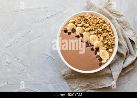 Frullato al cioccolato ciotola con le banane, granola e arachidi sulla luce di cemento grigio Sfondo. Una sana colazione vegetariana. Vista dall'alto. Foto Stock