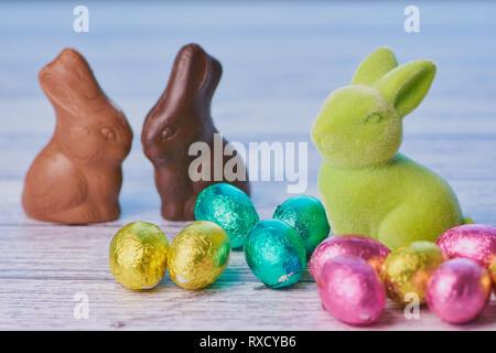 In primo piano un coniglietto di pasqua accanto avvolto lucido colorato di uova di cioccolato, sullo sfondo di due conigli di cioccolato non confezionato su bianco backg in legno Foto Stock