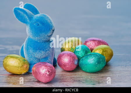 Blu coniglietto di pasqua con avvolto di uova di cioccolato contro il bianco sullo sfondo di legno con spazio di copia Foto Stock