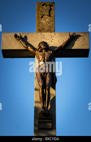 Bella croce di pietra crocifisso con Gesù Cristo scultura in bronzo colpito dalla luce del tramonto su un cielo blu. Foto Stock