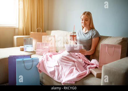 Shopping online a casa. Giovane donna bionda con il tè è guardando a tavoletta e la scelta di merci in negozio in linea mentre è seduto con coperta su un divano Foto Stock