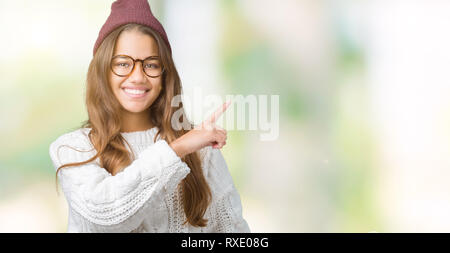 Giovane bella bruna hipster donna che indossa occhiali e cappello invernale su isolati di puntamento dello sfondo con la mano il dito a lato mostra pubblicizzare Foto Stock