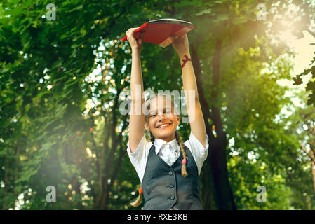 Studente schoolgirl felice con pig-tail in uniforme con libri in le mani sopra la testa in una luminosa giornata di sole Foto Stock