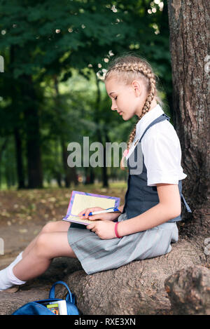 Ritratto di una studentessa adolescente in uniforme con uno zaino, in un parco nei pressi di un grande albero. schoolgirl la lettura di un libro Foto Stock