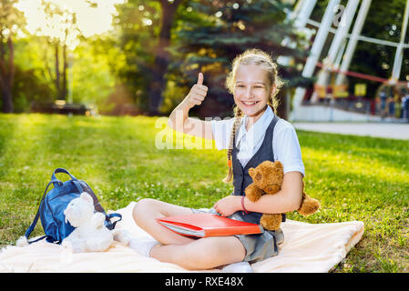 Schoolgirl studente sorridenti che mostra il pollice in alto si siede su una coperta nel parco in una giornata di sole. Il ragazzo è in possesso di un orso giocattolo in mano sua. c Foto Stock