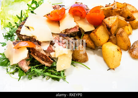 Cucina Italiana - Tagliata di Manzo (fette di angus roast beef) rustico con patate arrosto, parmigiano e insalata di rucola close up sulla piastra bianca Foto Stock