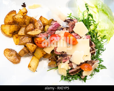 Cucina Italiana - vista superiore della Tagliata di Manzo (fette di angus roast beef) rustico con patate arrosto, parmigiano e insalata di rucola su bianco pla Foto Stock